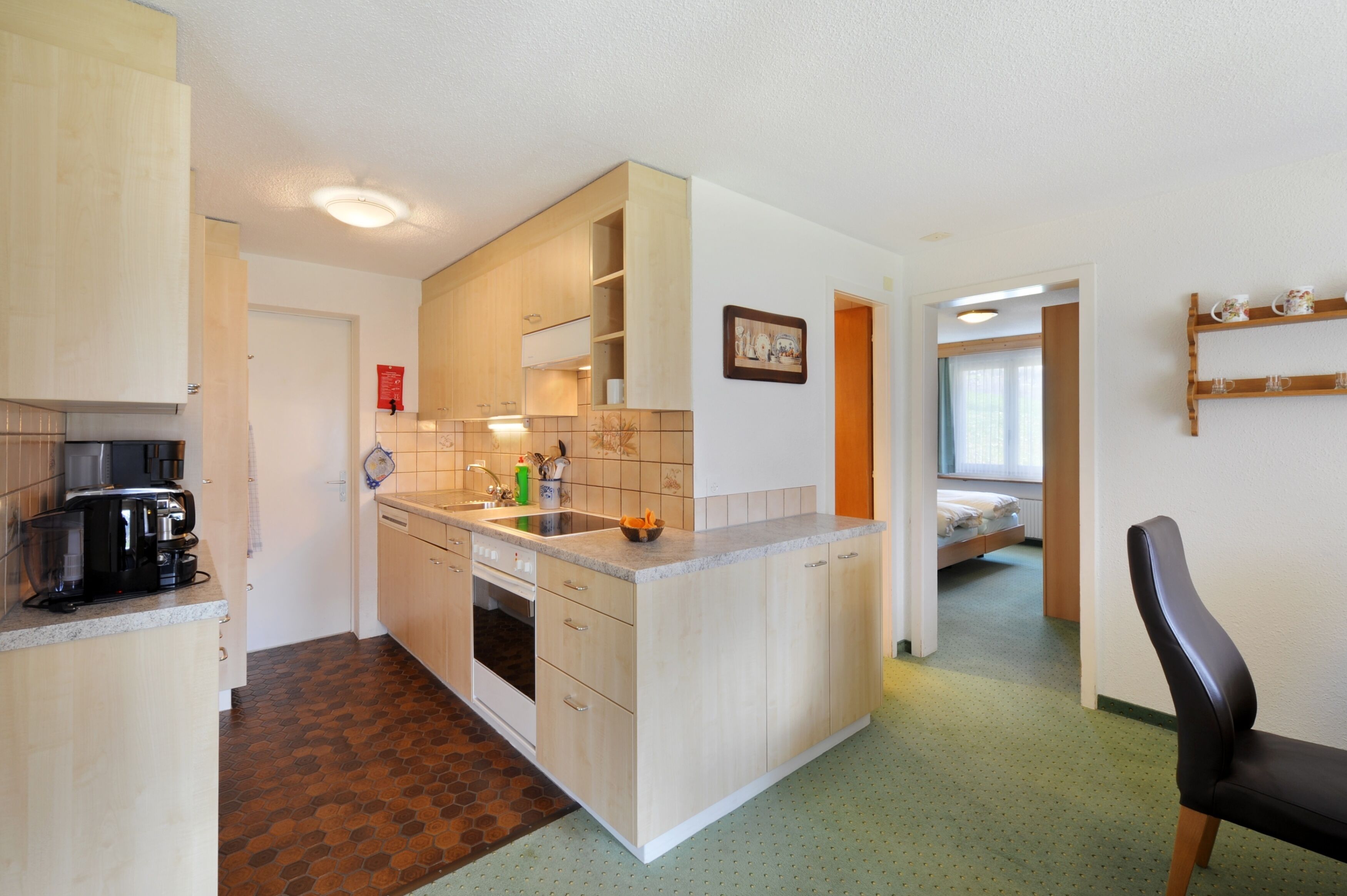 A compact kitchen with a coffee maker, sink, and cabinets. A bed is visible in the adjacent room.