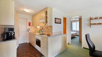 A compact kitchen with a coffee maker, sink, and cabinets. A bed is visible in the adjacent room.