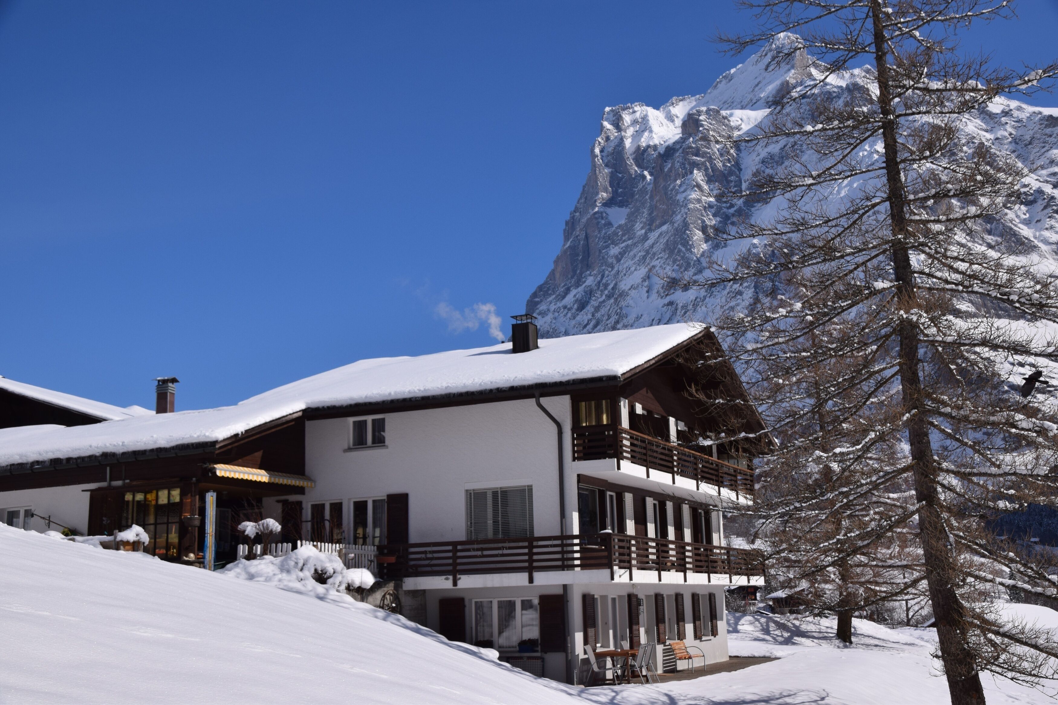 A two-story chalet with a balcony, surrounded by snow-covered landscape and a mountain in the background.