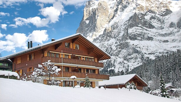 Een houten chalet met meerdere balkons, omringd door besneeuwde bergen en bomen.
