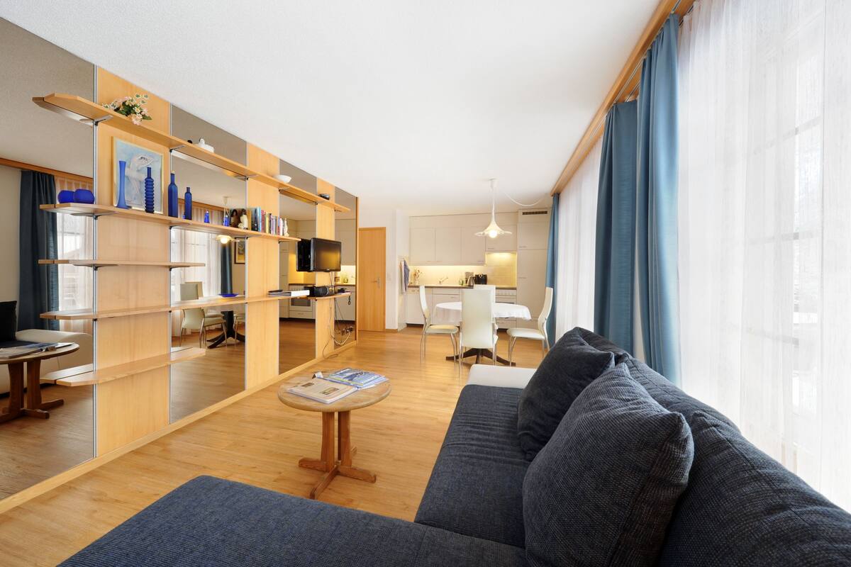 Una sala de estar moderna con un sofá, una mesa de centro y un comedor con una mesa y sillas blancas. Se observa una cocina con estufa y armarios.