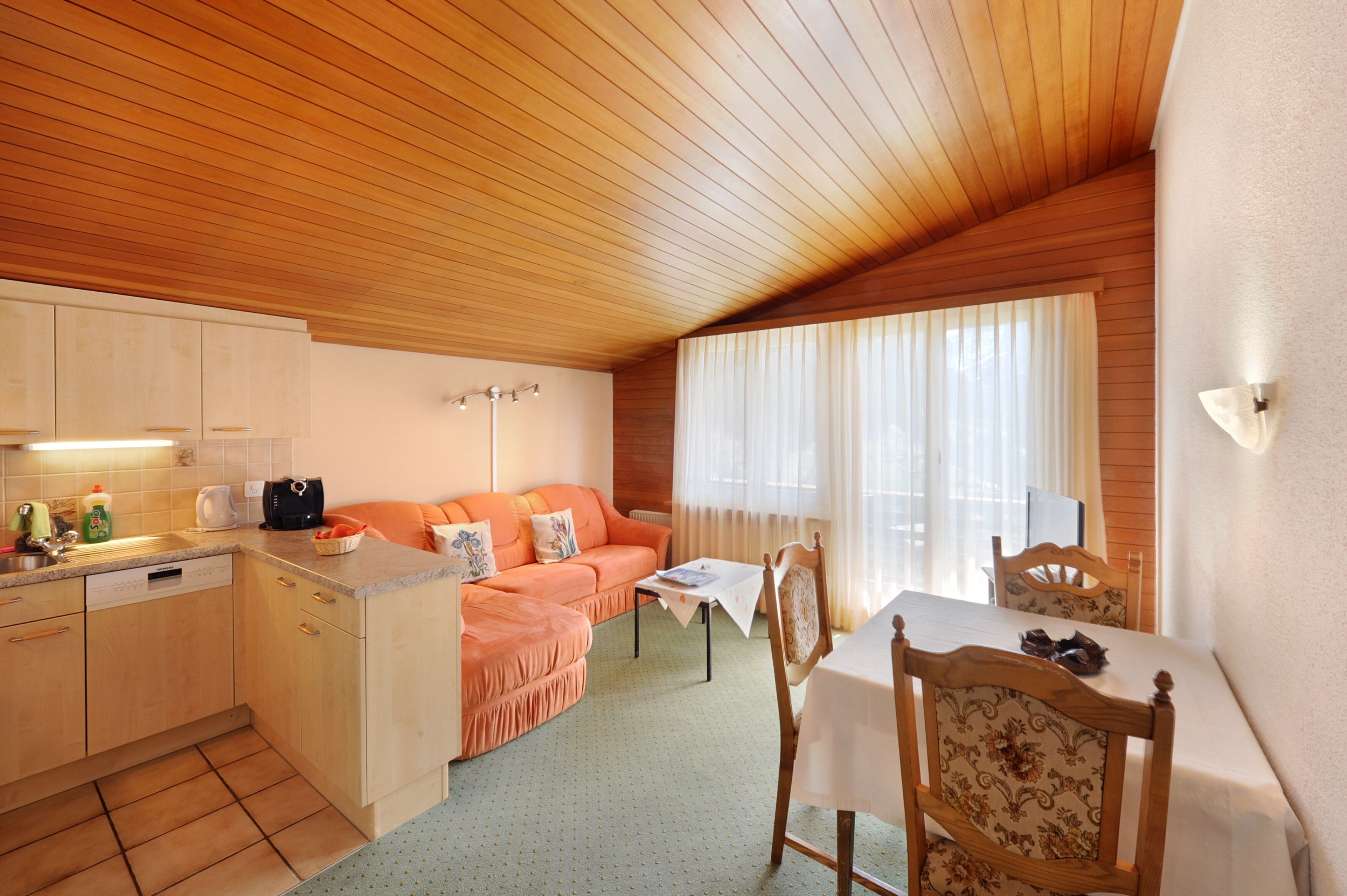Une cuisine compacte avec des meubles en bois, un évier et un plan de travail. On y trouve un coin repas avec une table et des chaises, ainsi qu’un canapé dans l’espace de vie.