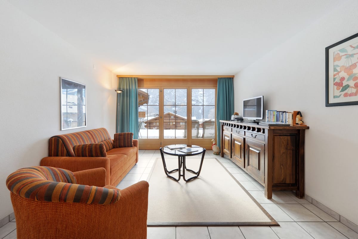 Una sala de estar con un sofá, un sillón y una mesa de centro de vidrio. Hay una zona de cocina con armarios y una estufa.