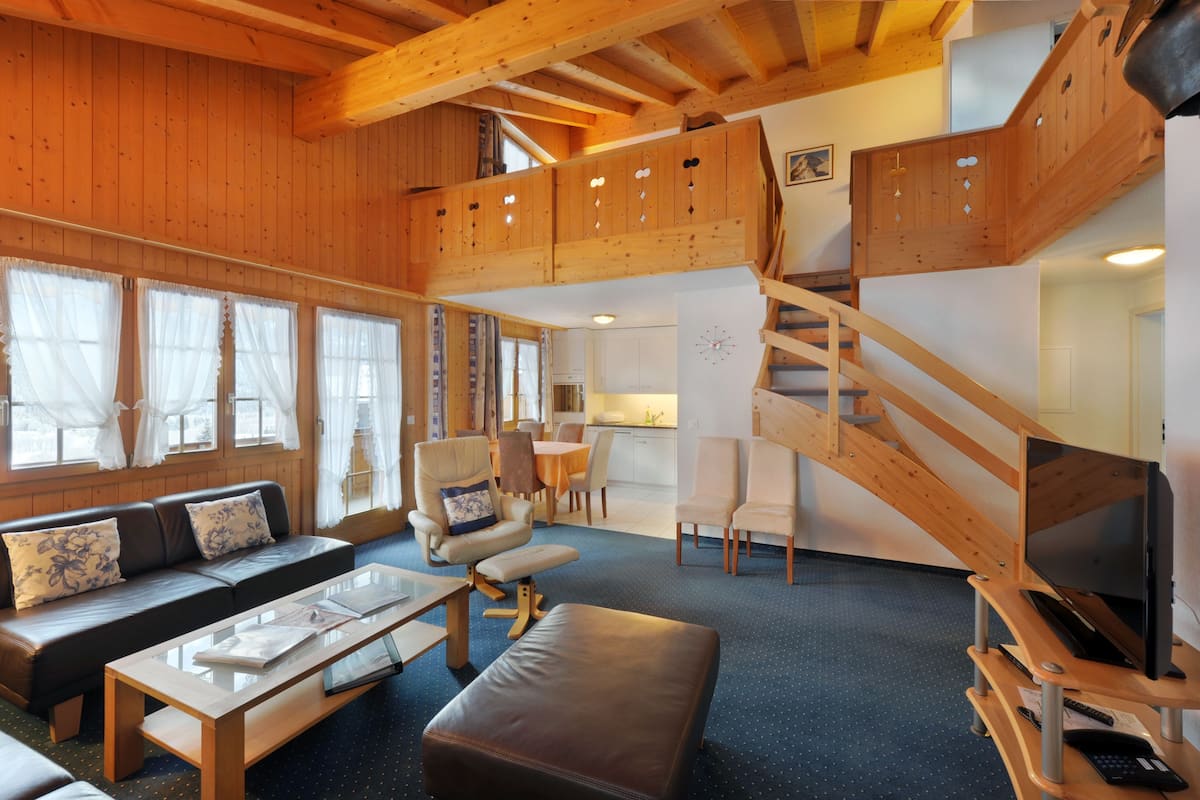Amplia sala de estar con una escalera de madera, un conjunto de sofás, una mesa de centro y un televisor sobre un mueble de madera.