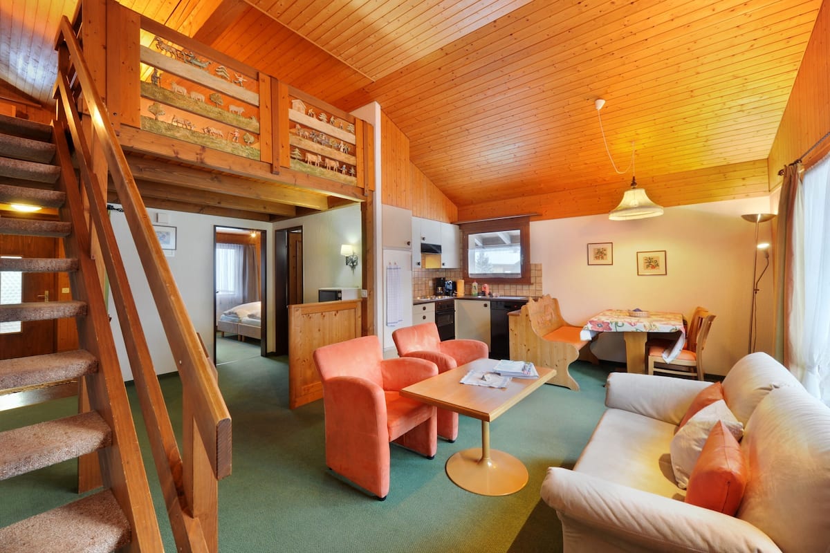 Un acogedor interior de cabaña con una escalera de madera, un comedor con mesa y sillas, un sofá y una zona de cocina.