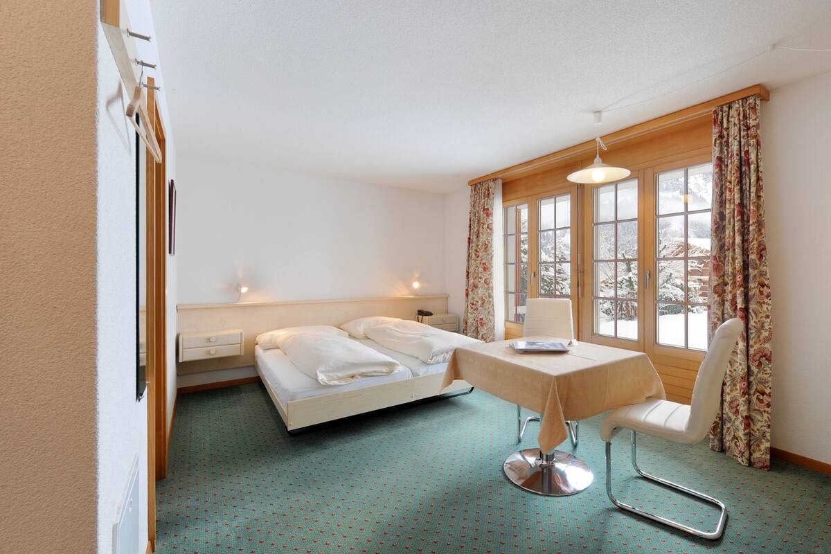 Habitación de hotel con cama, una mesa, una silla y ventanas amplias.