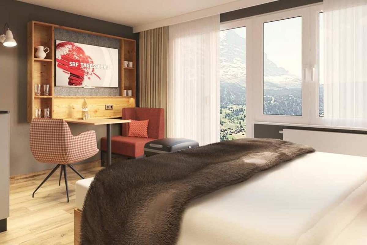 Habitación de hotel con cama, escritorio, silla, televisor y ventana con vista a la montaña.