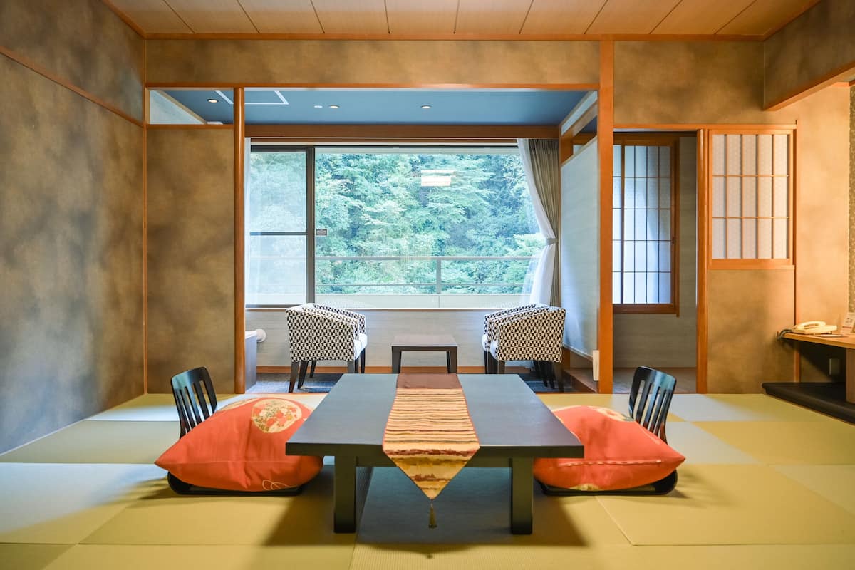 A traditional Japanese room with tatami flooring, a low table with cushions, and a large window offering a forest view.