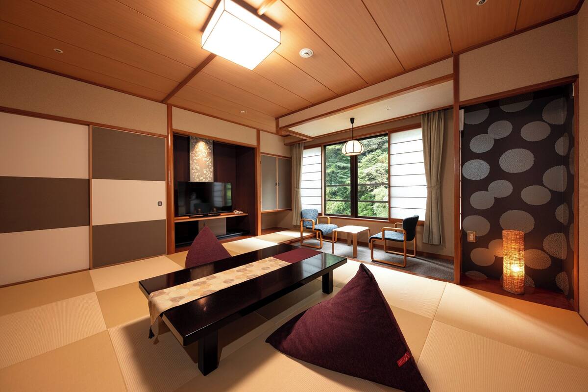 A traditional Japanese room with tatami flooring, a low table, cushions, a television, and a view of greenery through the window.