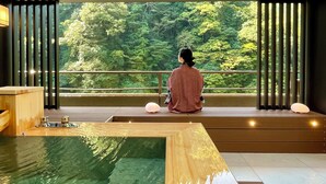 A person sitting by a window overlooking a forest, with a wooden table and a small water feature in the foreground. - Tsuetate Onsen Hizenya (Oguni)