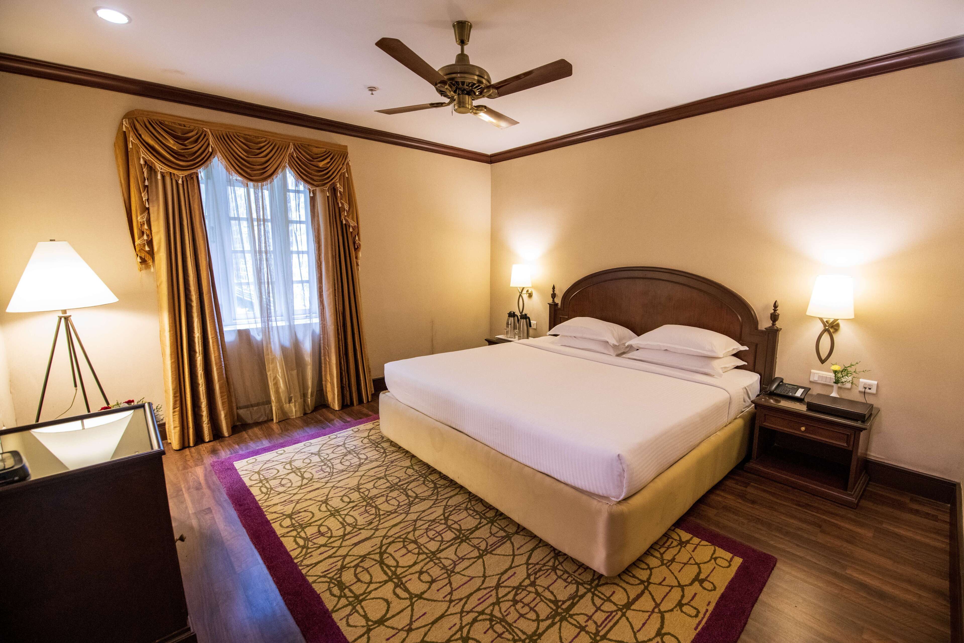 Quarto de hotel com uma cama grande, ventoinha no teto, mesa com luminária e janela com cortinas.