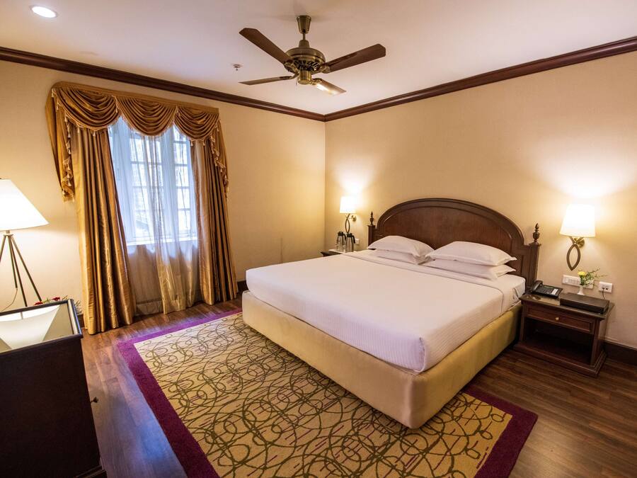 Quarto de hotel com uma cama grande, ventoinha no teto, mesa com luminária e janela com cortinas.