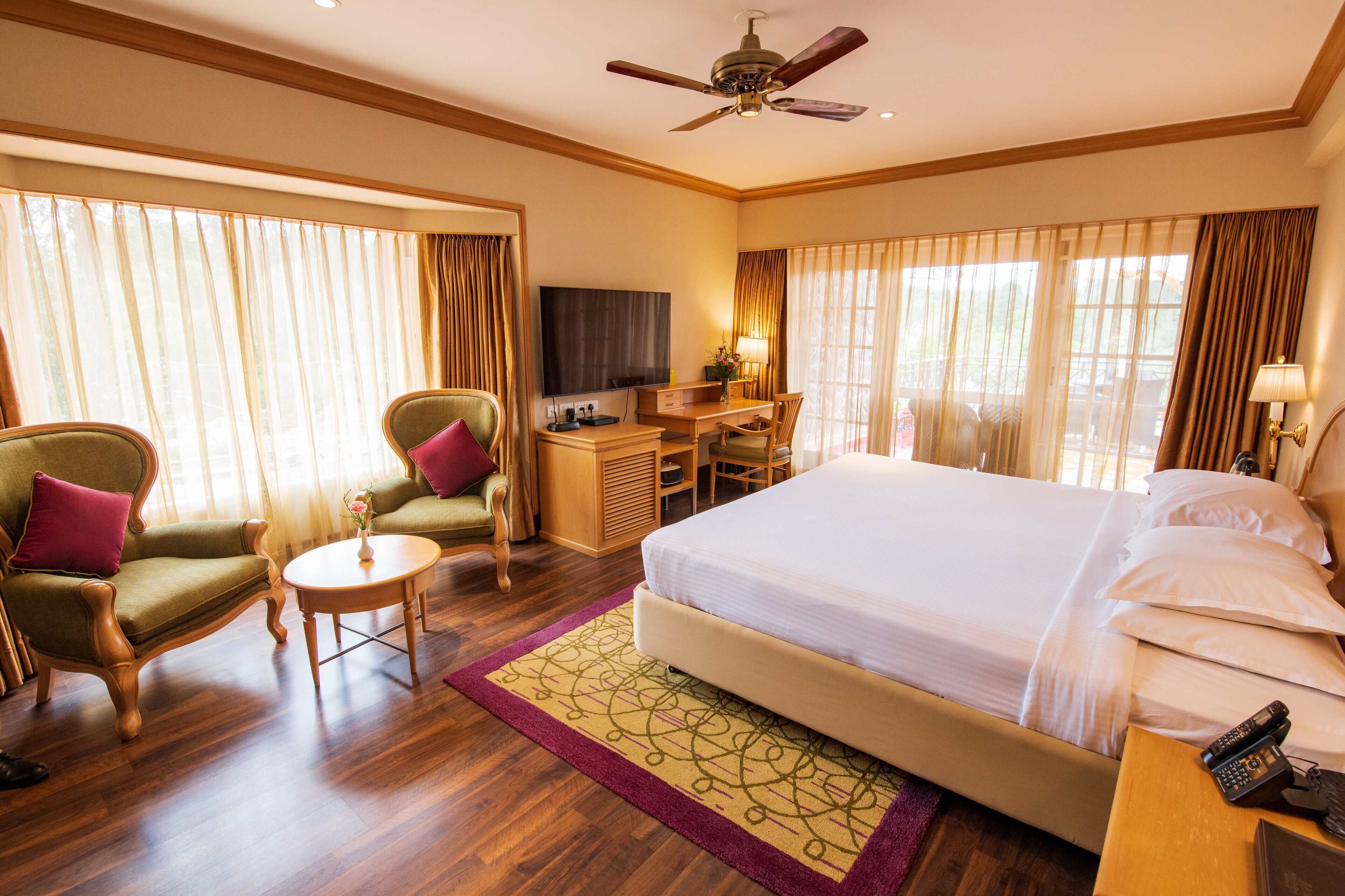 A hotel room with a large bed, two armchairs, a desk with a chair, a television, and a ceiling fan.