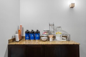 A kitchen counter with hot water dispensers, a medium roast coffee dispenser, a water cooler, a glass pitcher with lemon slices, and a metal rack with utensils. - Comfort Inn & Suites Scott-West Lafayette (Scott)