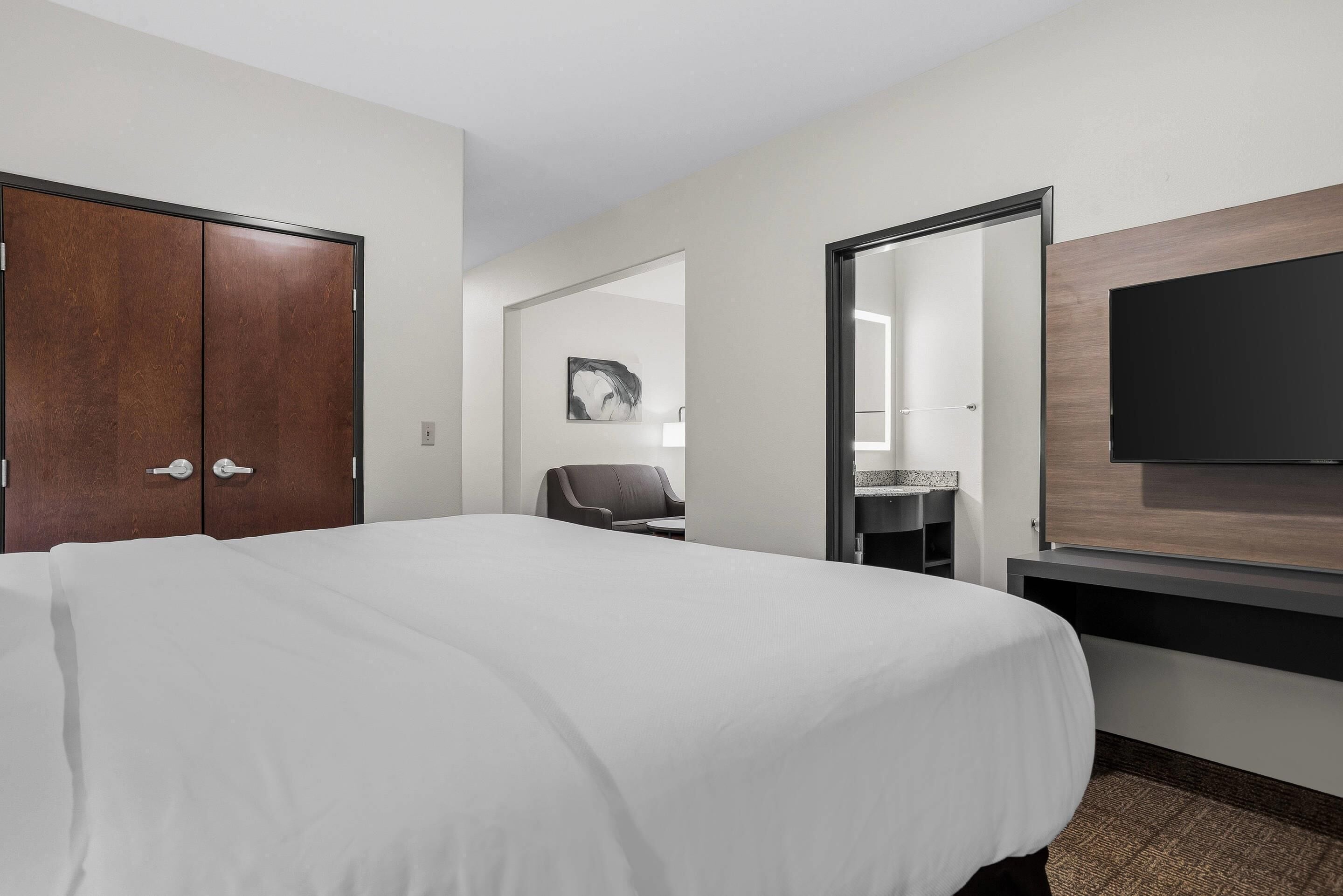 A hotel room with a large bed, a desk, a chair, a flat-screen TV mounted on the wall, and a door leading to another room.