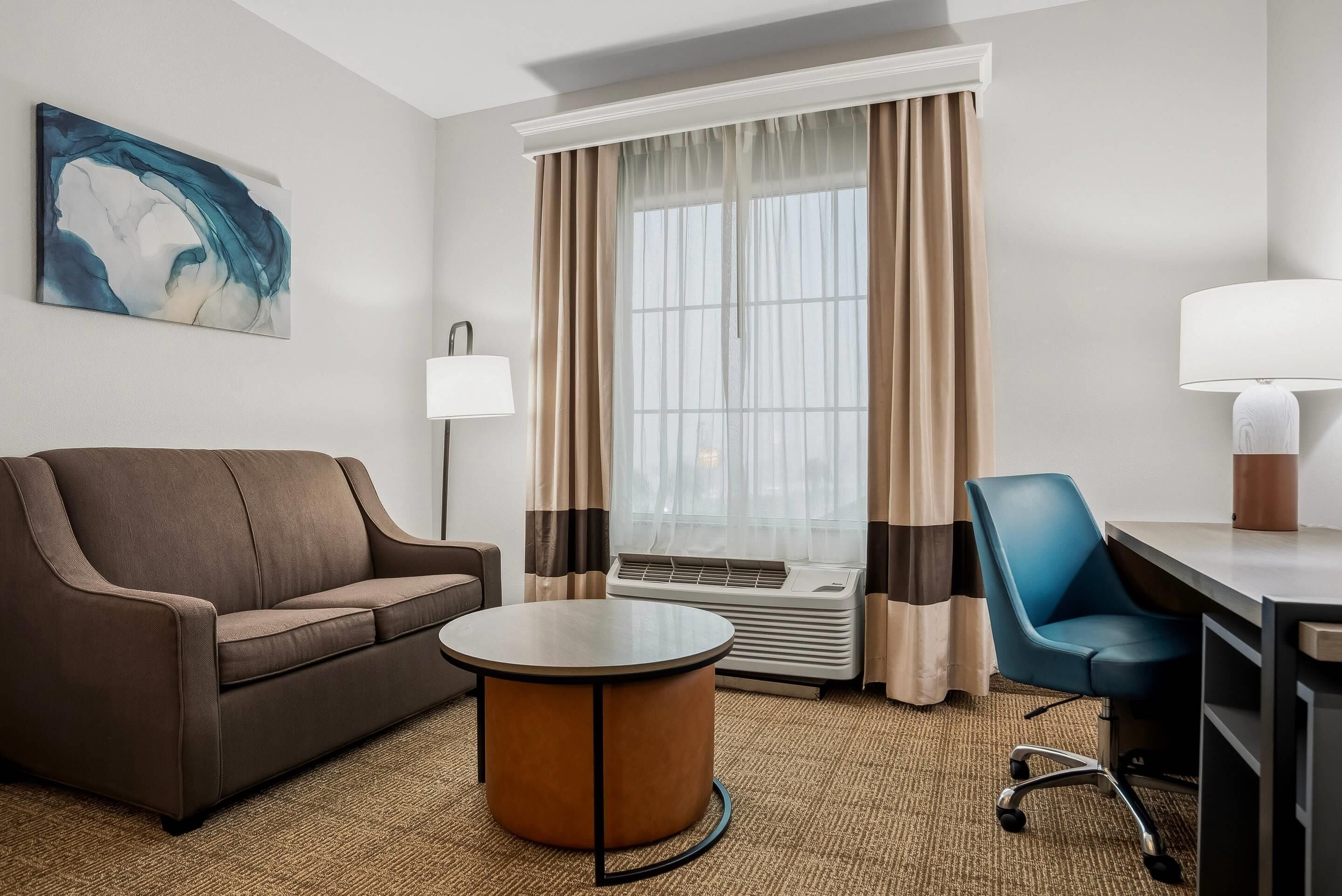 Habitación de hotel con sofá, una mesita redonda, escritorio con silla, ventana con cortinas y una lámpara de pared.