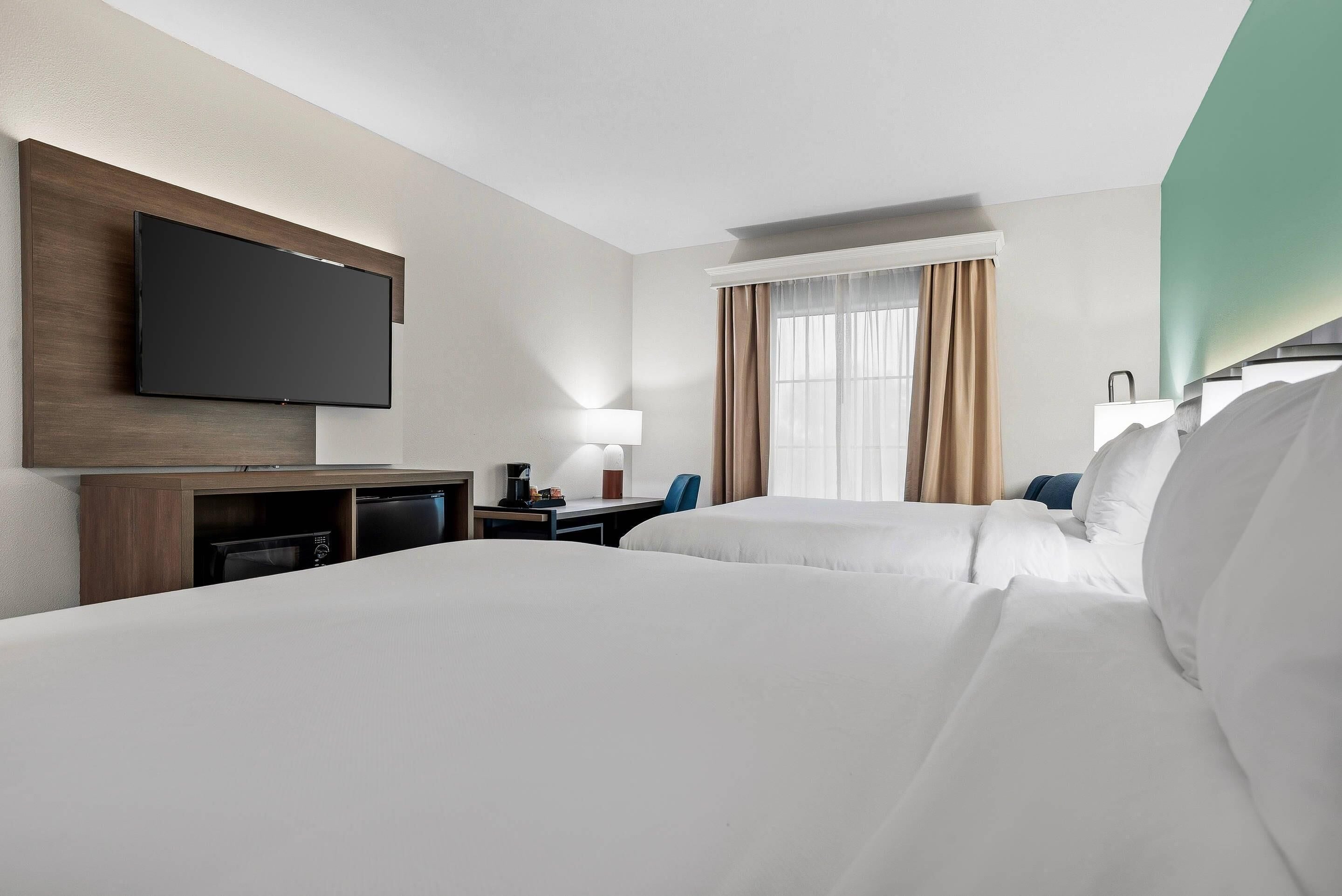 Habitación de hotel con televisor de pantalla plana en la pared, un escritorio con lámpara, una silla y una cama con ropa blanca.