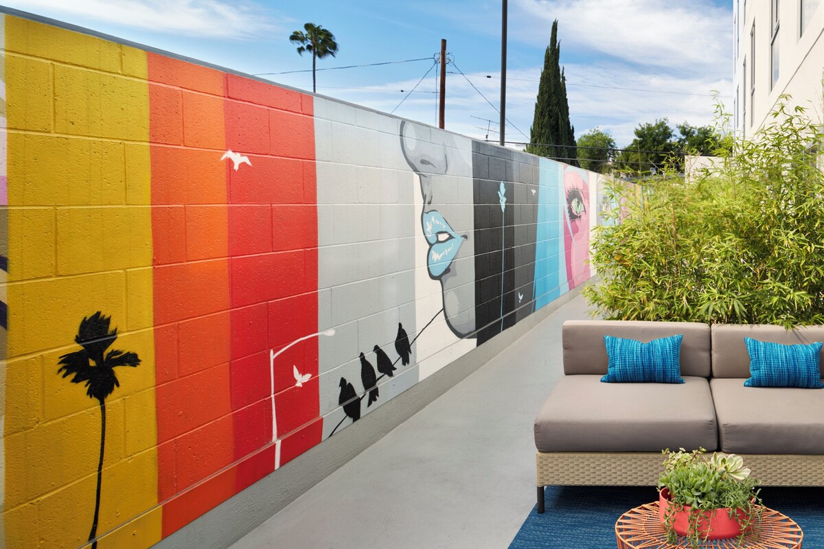 A colorful mural on a wall, a gray sofa with blue cushions, and a potted plant on a blue mat.
