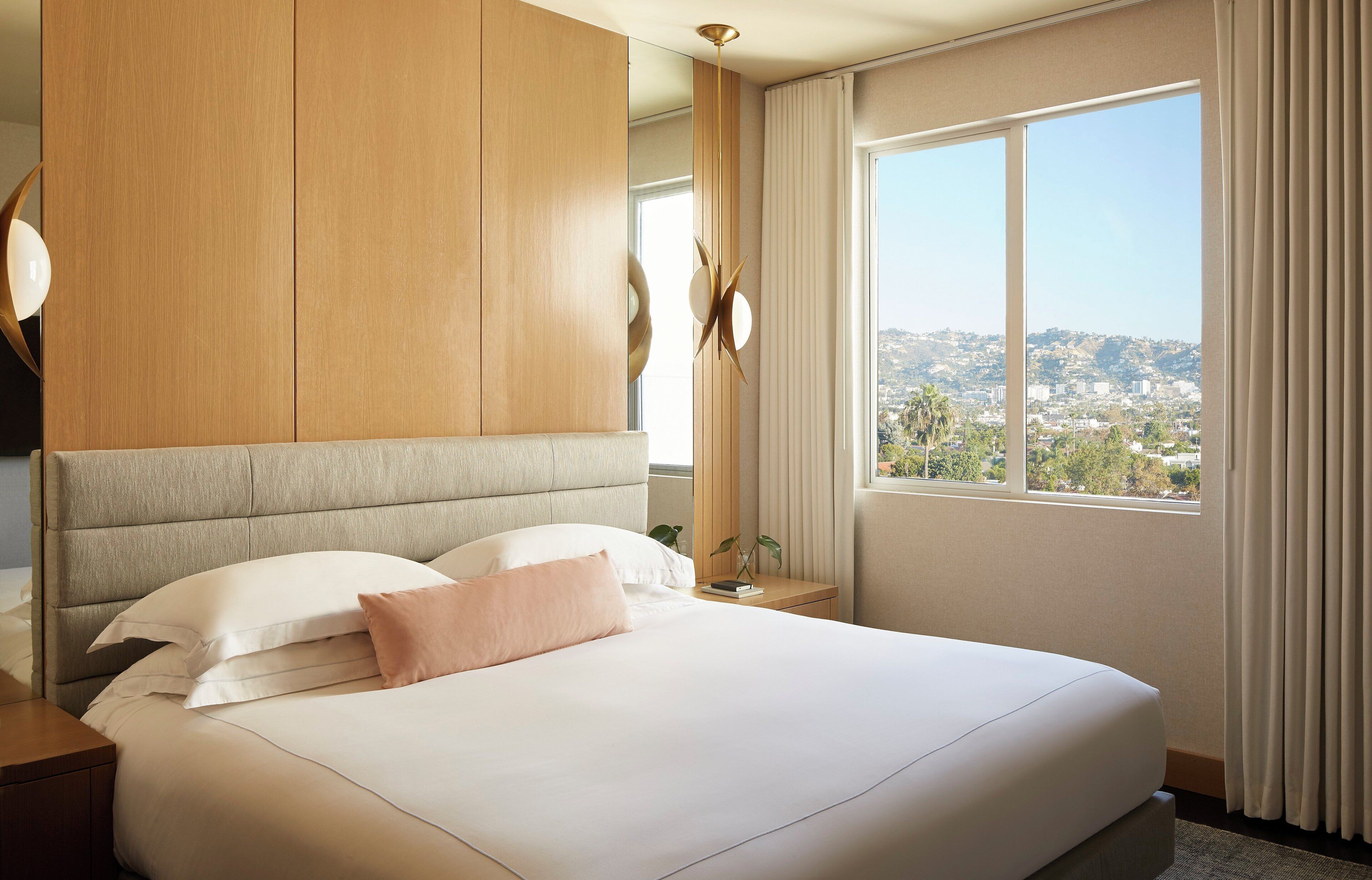 A hotel room with a large bed, a wooden headboard, a nightstand, a bedside table with a lamp, and a window with a view of a cityscape.