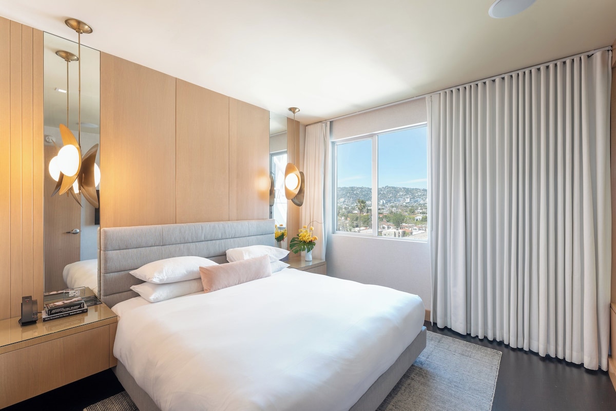 Habitación de hotel con una cama grande, una mesita de noche, un espejo y vistas a un paisaje urbano por la ventana.