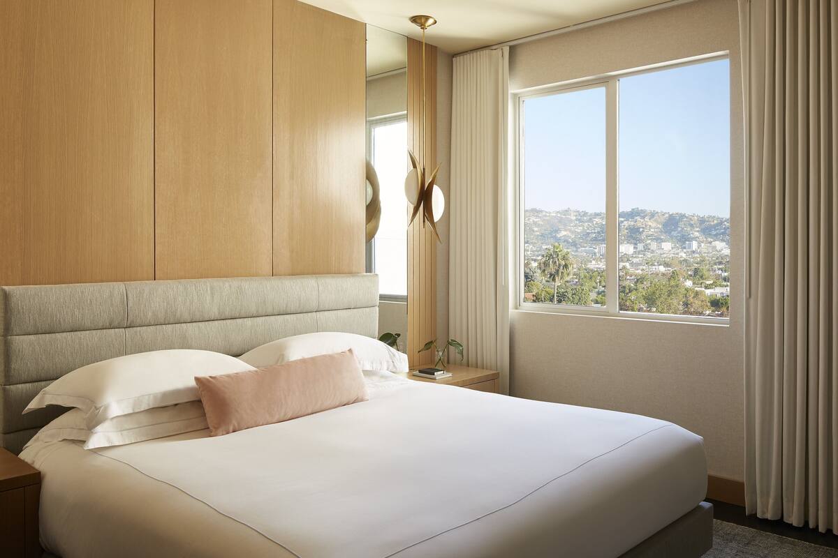 Een moderne hotelkamer met een groot bed, een houten hoofdbord, uitzicht op een stadsbeeld en een groot raam met gordijnen.