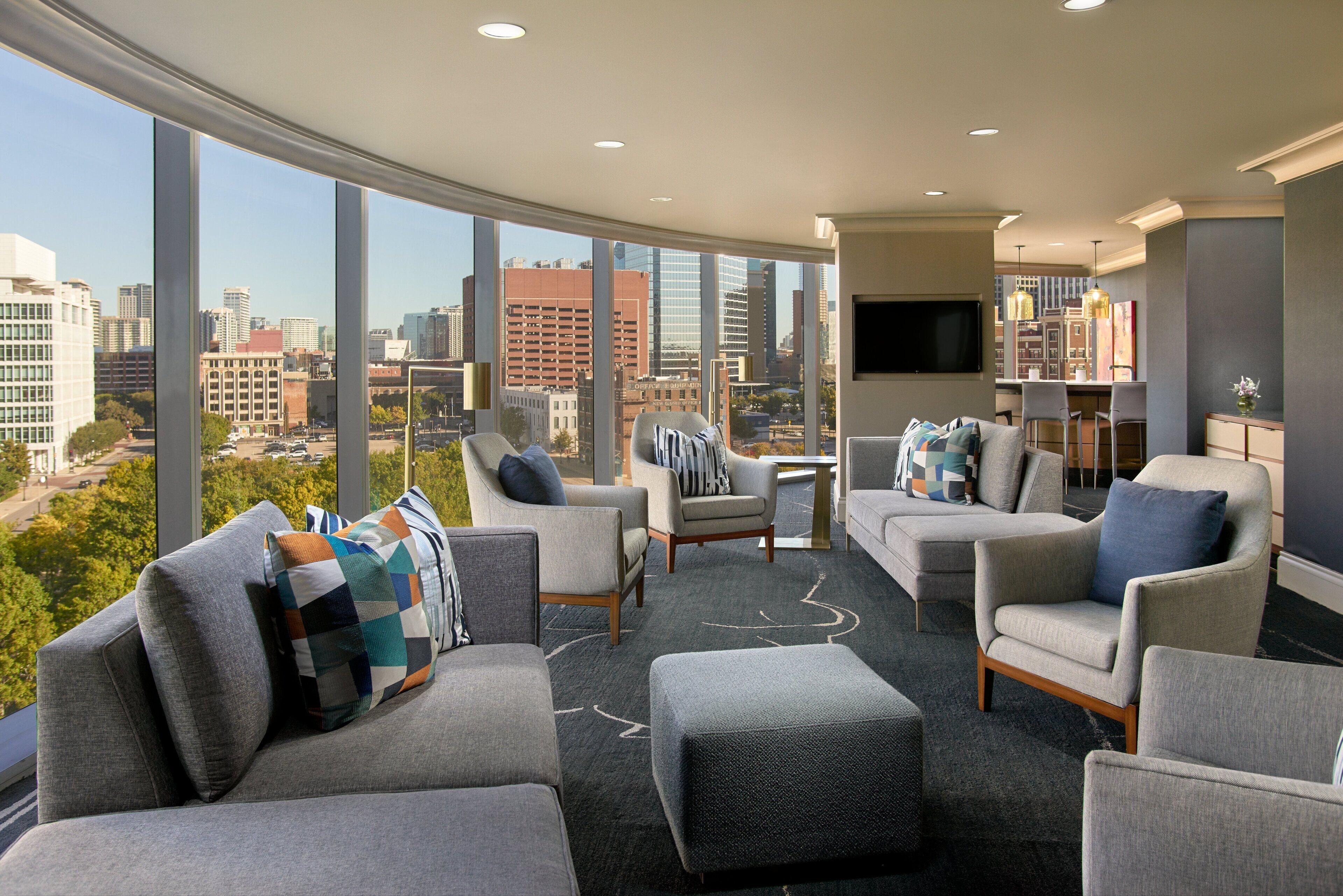 Ein modernes Wohnzimmer mit Blick auf die Stadt, ausgestattet mit einer Couch, Sesseln, einem Couchtisch und einem an der Wand befestigten Fernseher.