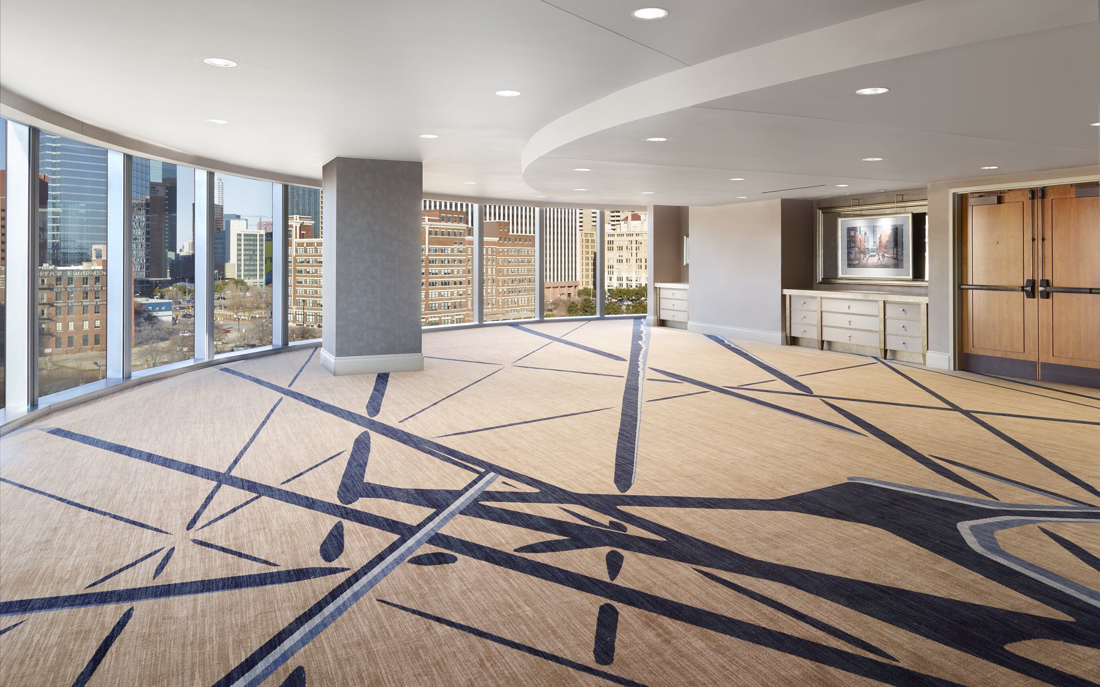A spacious lobby with large windows offering city views, a modern ceiling design, and a carpet with geometric patterns.