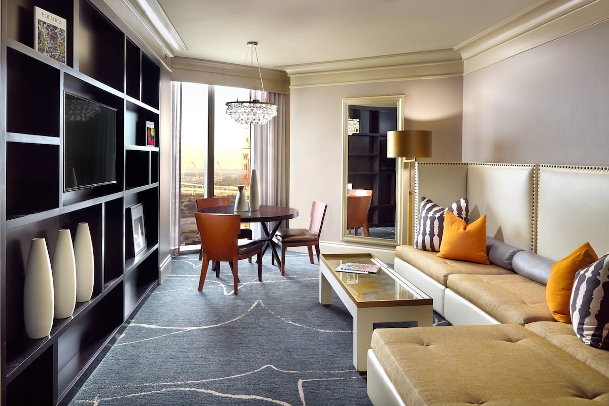 Ein modernes Hotelzimmer mit großem Fenster, einer Couch, einem Couchtisch, einem Bücherregal und einem Essbereich mit Stühlen.