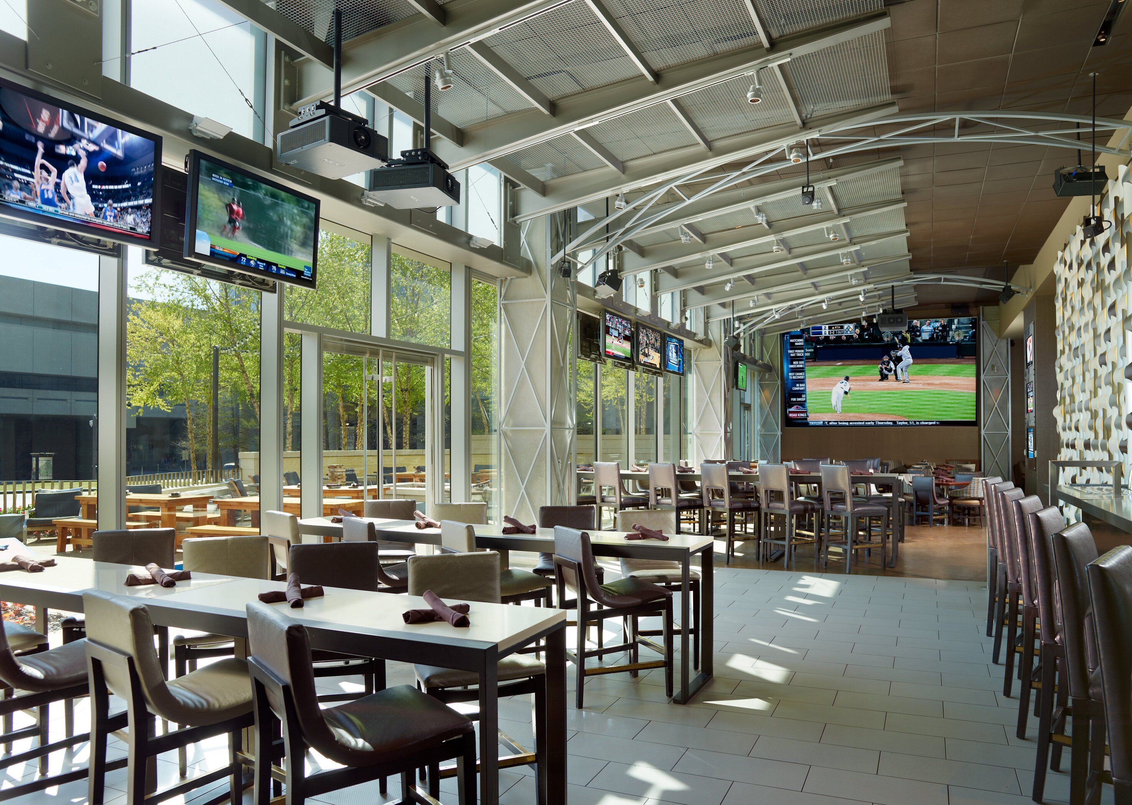 A spacious indoor dining area with multiple tables and chairs, large windows, and multiple screens displaying sports events.