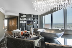 A modern hotel room with a large dining table, a chandelier, a city view, and a bookshelf with decorative items.