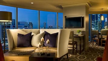 A modern hotel room with a large window offering a city view, a sofa with decorative pillows, a small table with candles, and a flat-screen TV mounted on the wall.