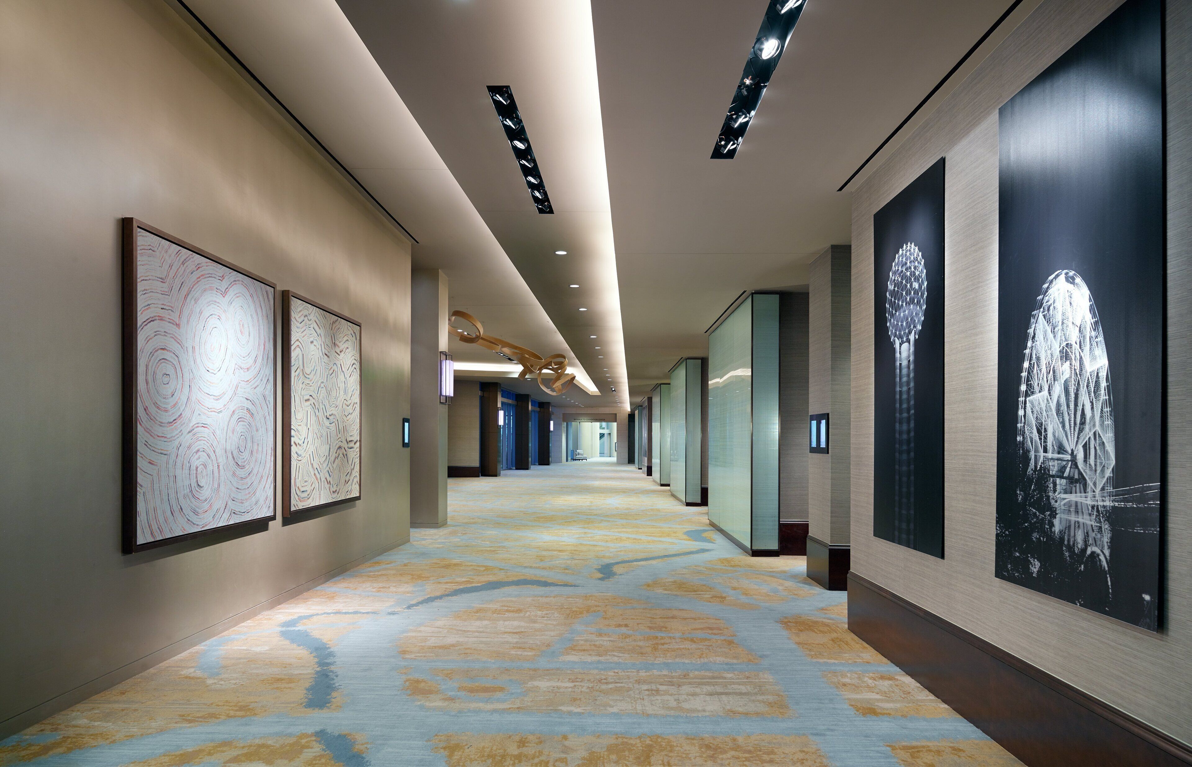 A long, well-lit hallway with patterned floor tiles, framed artworks on the walls, and recessed lighting.