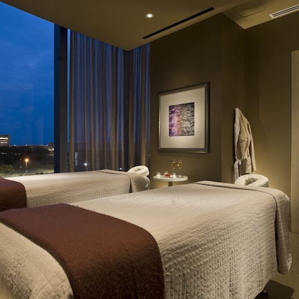 A hotel room with two beds, a small table with a drink, a framed picture on the wall, and a view of the city at dusk.