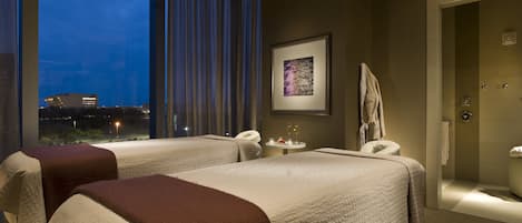 A hotel room with two beds, a small table with a drink, a framed picture on the wall, and a view of the city at dusk.