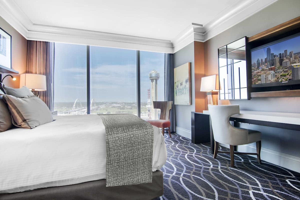 Ein Hotelzimmer mit einem großen Bett, einem Schreibtisch, einem Sessel und Blick auf die Stadt durch große Fenster.