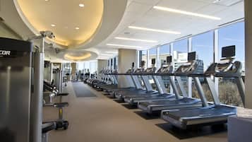 Un gimnasio con cintas de correr alineadas a lo largo de una pared con ventanas, iluminación empotrada en el techo y equipo de ejercicio a la izquierda.