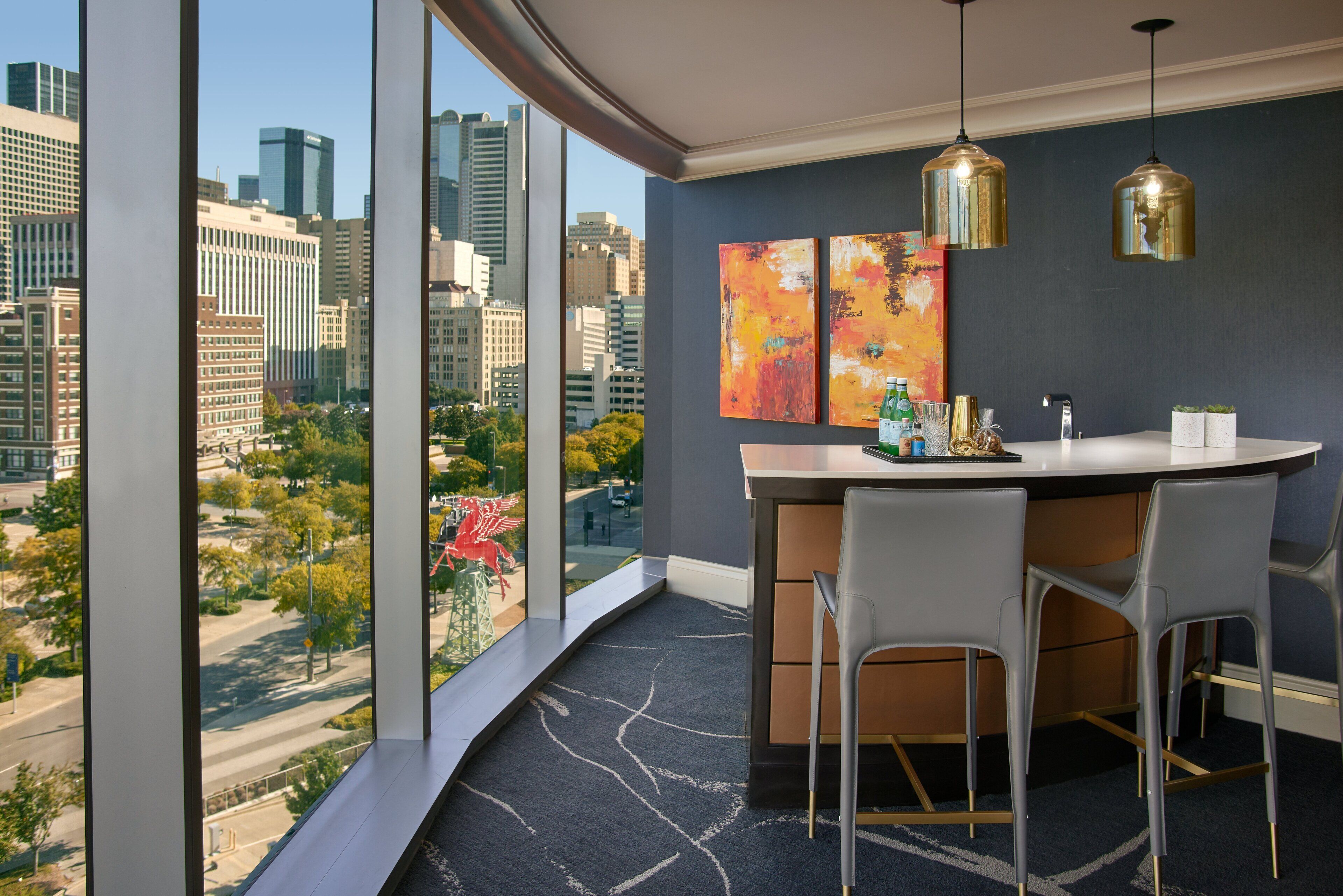 A modern hotel room with a city view, a curved desk with two chairs, and two abstract paintings on the wall.