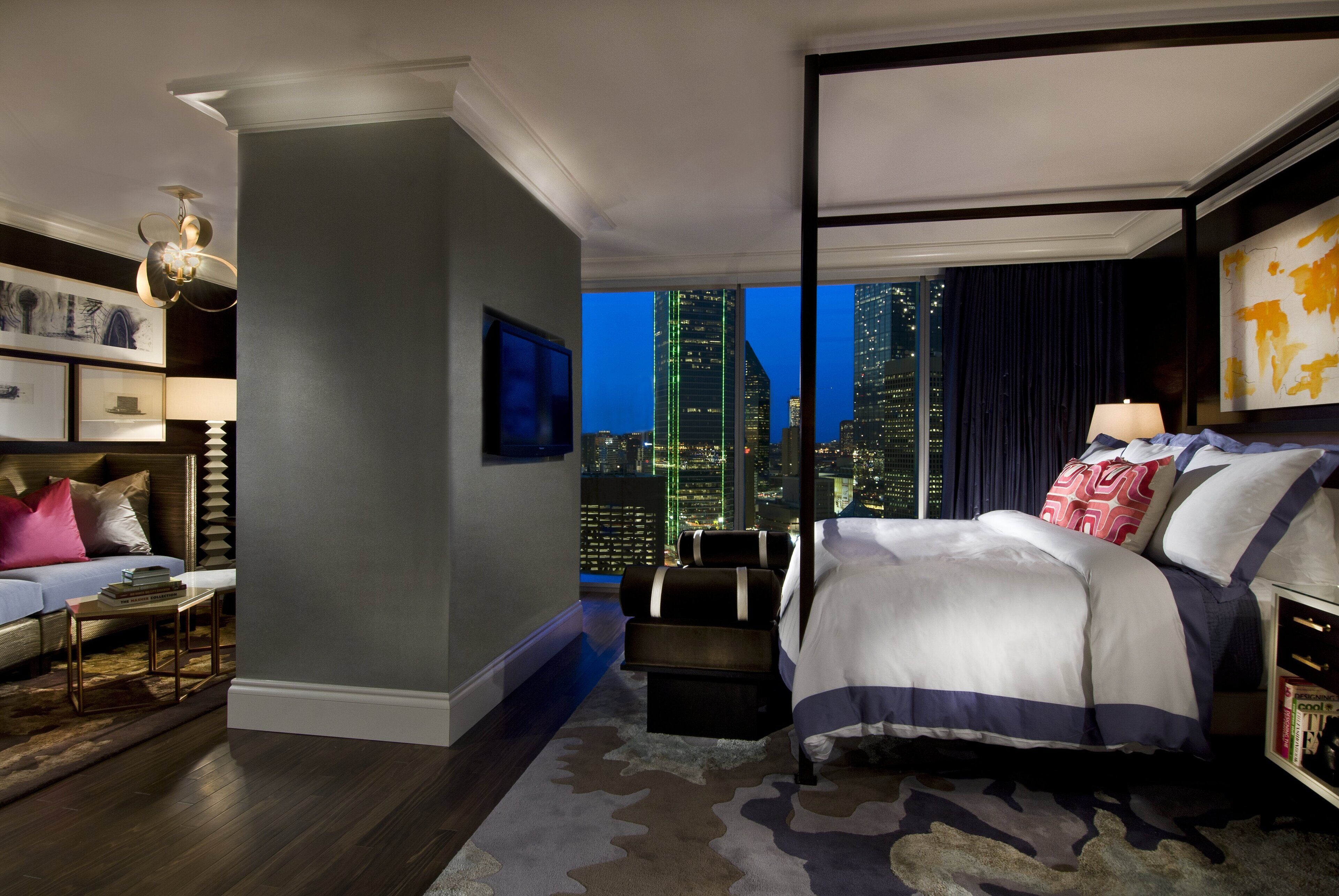 A modern hotel room with a city view, a large bed, a seating area, and a bookshelf.