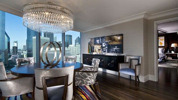 A modern living room with a city view, a chandelier, a dining area with chairs, a sofa, a TV, and a cabinet. - Omni Dallas Hotel (Dallas)