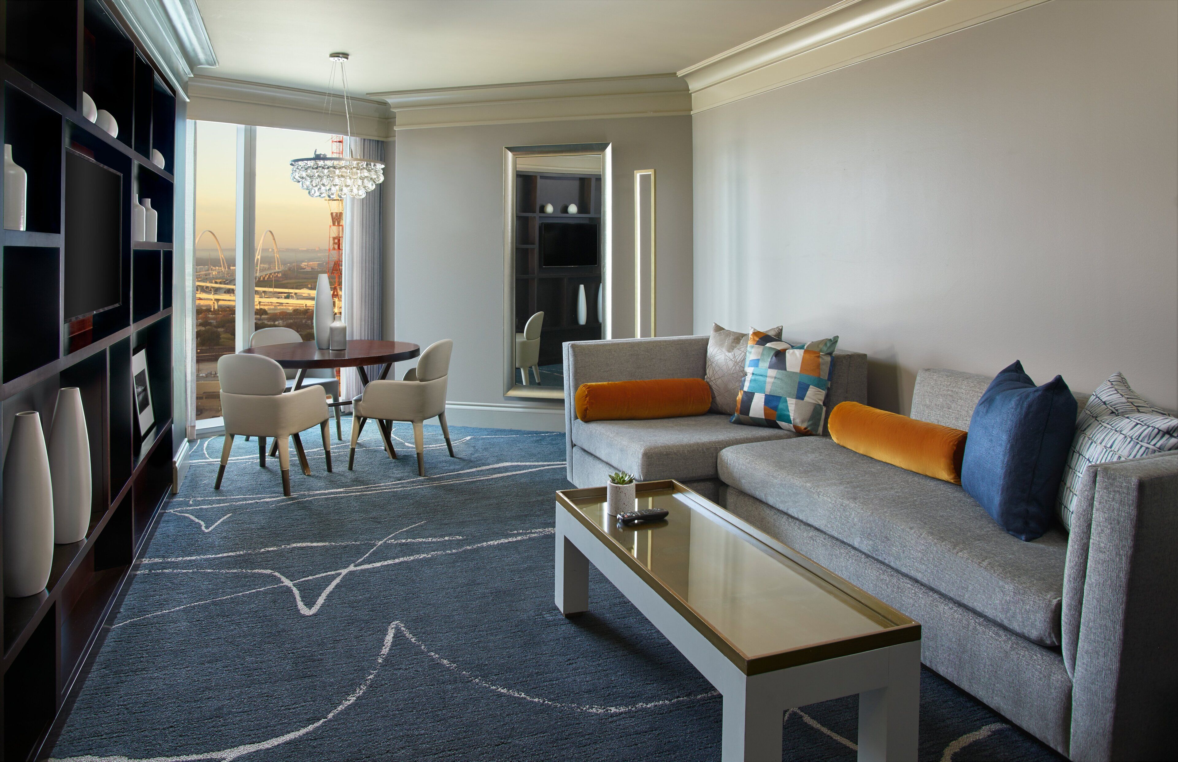 A modern living room with a sofa, coffee table, and bookshelf. There is a dining area with chairs and a table. The room has large windows with a view of a bridge.