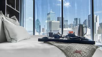 Quarto de hotel com uma cama, almofadas, um tabuleiro com uma cafeteira e vista para o horizonte urbano através de amplas janelas.