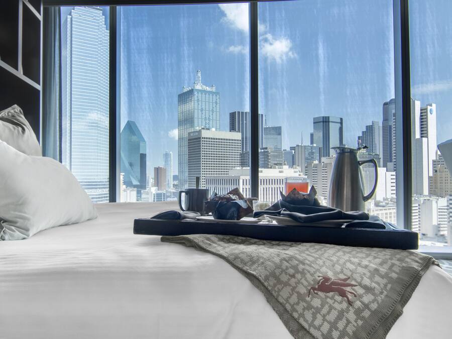 Quarto de hotel com uma cama, almofadas, um tabuleiro com uma cafeteira e vista para o horizonte urbano através de amplas janelas.