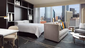 Habitación de hotel moderna con cama, sofá y zona de comedor, que ofrece vistas a un horizonte urbano.