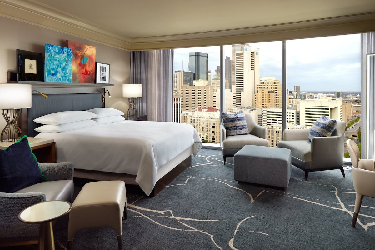 Ein Hotelzimmer mit einem großen Bett, einer Sitzecke mit Sesseln und Hocker, Blick auf die Stadt durch große Fenster und Kunstwerken an der Wand.