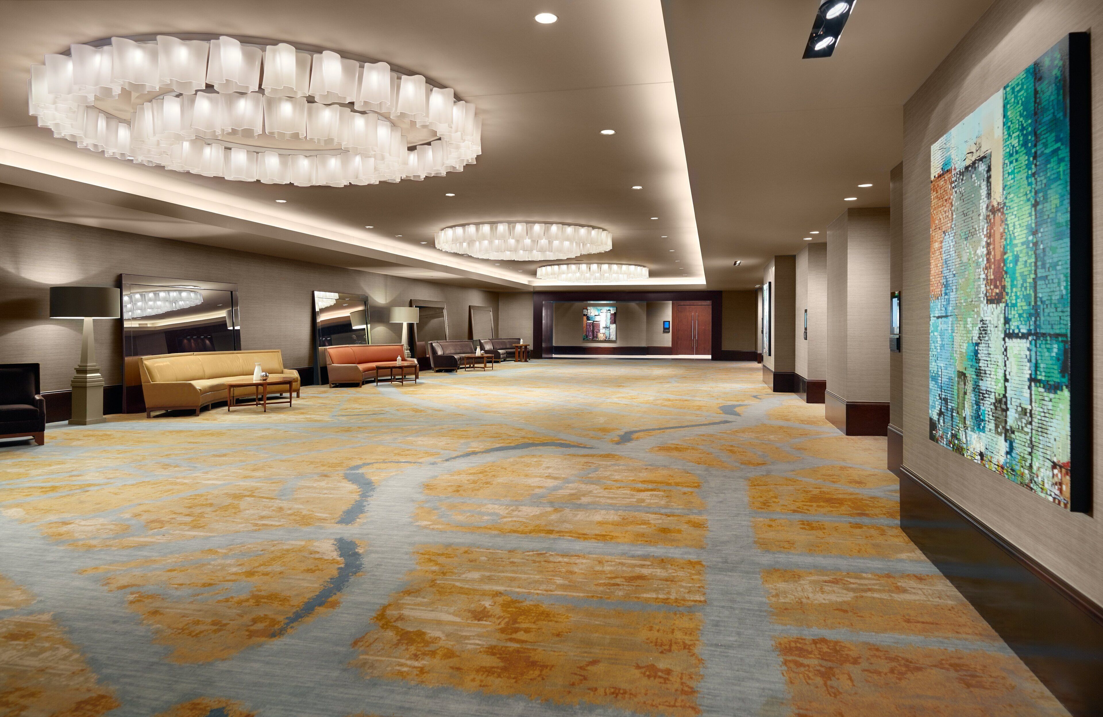 A spacious hotel lobby with a large carpeted area, multiple seating arrangements, and a prominent art piece on the wall.