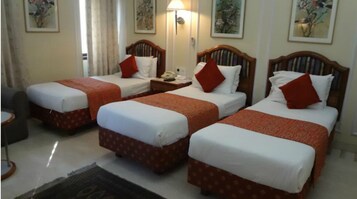 A hotel room with three single beds, each with a wooden headboard, white bedding, and red accents. There is a bedside table with a lamp, a telephone, and a small table with a chair. The room has a large window with curtains, and artwork is displayed on the walls.