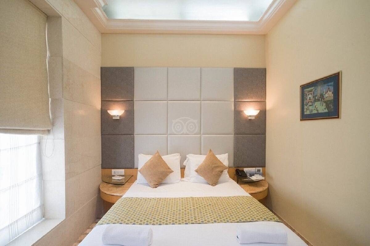 A hotel room with a large bed, two bedside tables, a nightstand, a window with blinds, and a ceiling light.