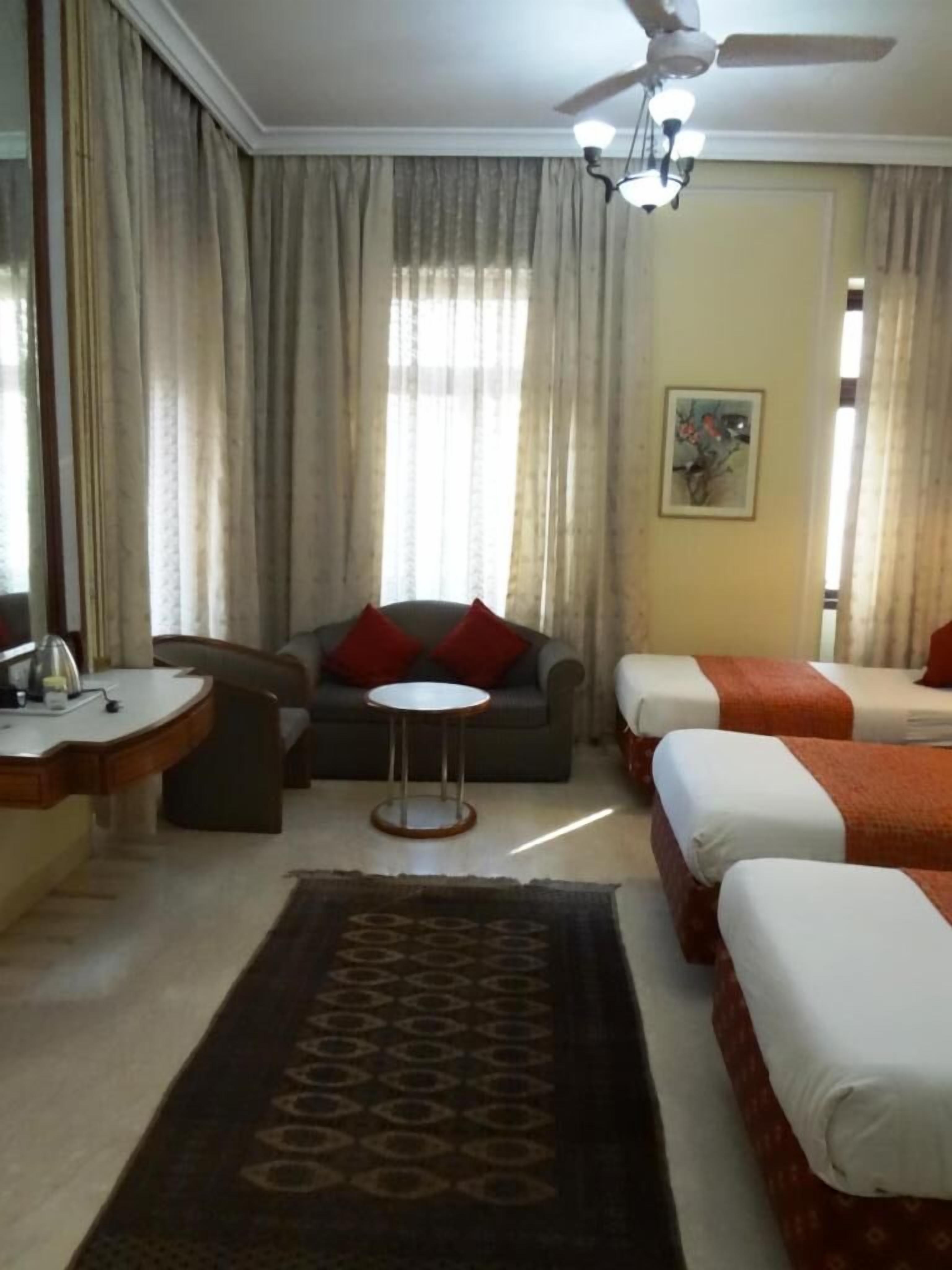 Habitación de hotel con sofá, dos camas individuales, una mesita, ventilador de techo y un ventanal grande con cortinas.