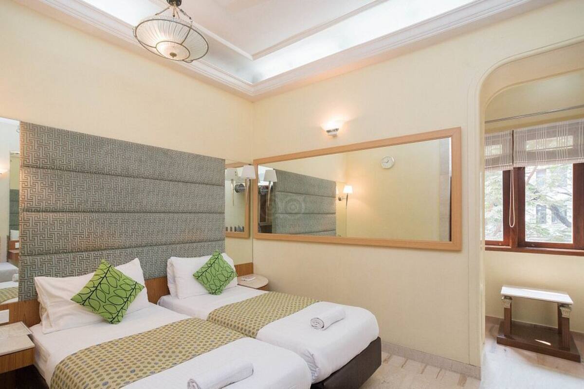 A hotel room with two beds, a large mirror, a bedside table, a small bench, and a ceiling light.