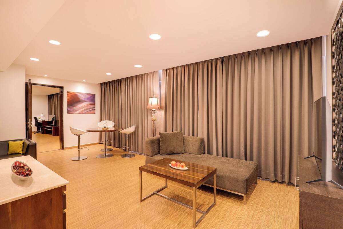 Uma sala de estar moderna com sofá, mesa de centro, área de refeições com abajur e vista para o horizonte urbano através de grandes janelas.